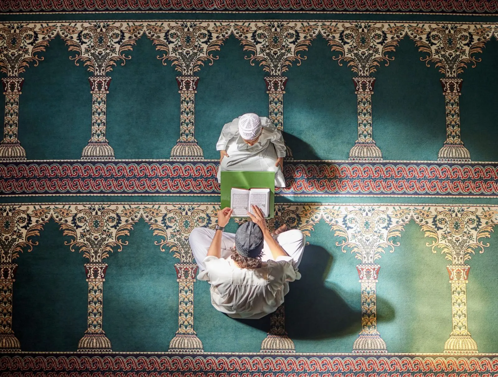 Two people reading the Qur’an together on a patterned prayer carpet